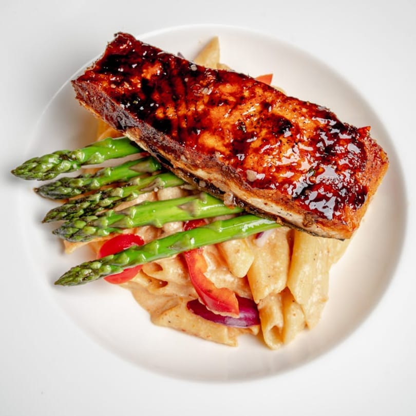 Glazed salmon fillet with asparagus spears and penne pasta with red peppers on a white plate.