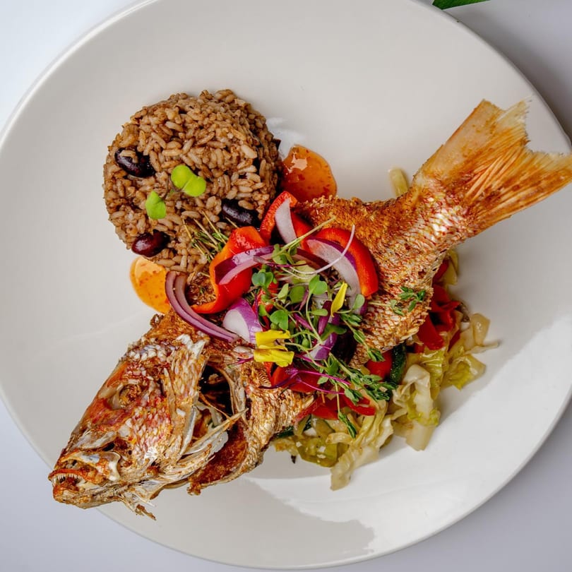 Whole fried Caribbean snapper with rice and peas, topped with colorful vegetables and microgreens.