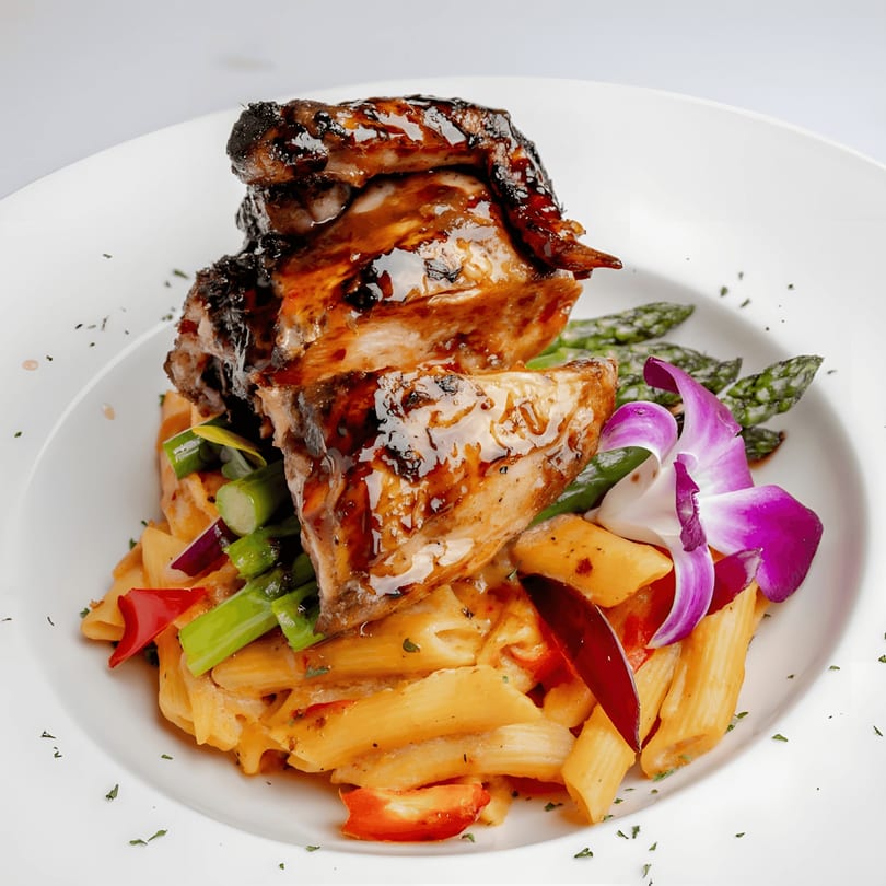 Glazed Caribbean jerk chicken with penne pasta, asparagus, bell peppers, and an orchid garnish.
