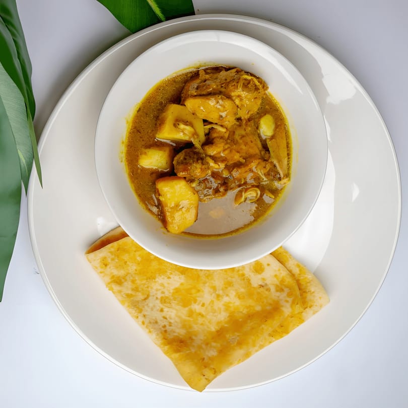 Caribbean chicken curry with potatoes in a white bowl, served with a folded roti on a white plate.