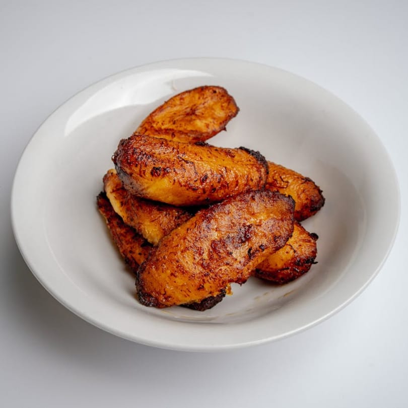 A white bowl filled with golden-brown, fried Caribbean plantains, glistening with oil.