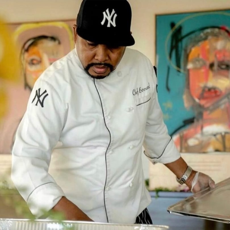 Chef Bernard, wearing a Yankees cap and chef's coat, prepares Caribbean food in a serving tray.