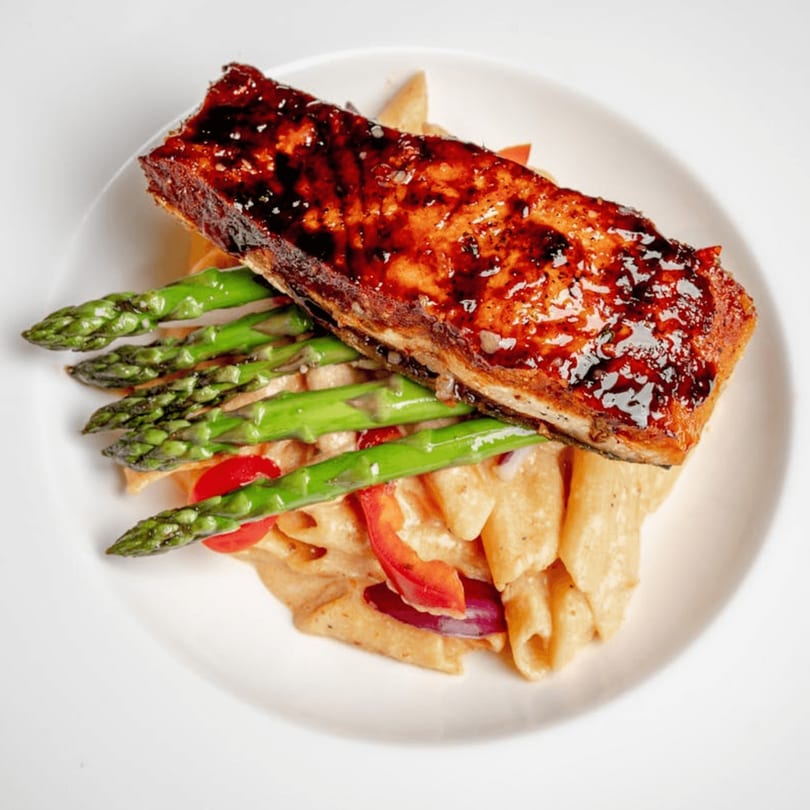 Glazed salmon fillet over penne pasta with red peppers, onions, and asparagus on a white plate.