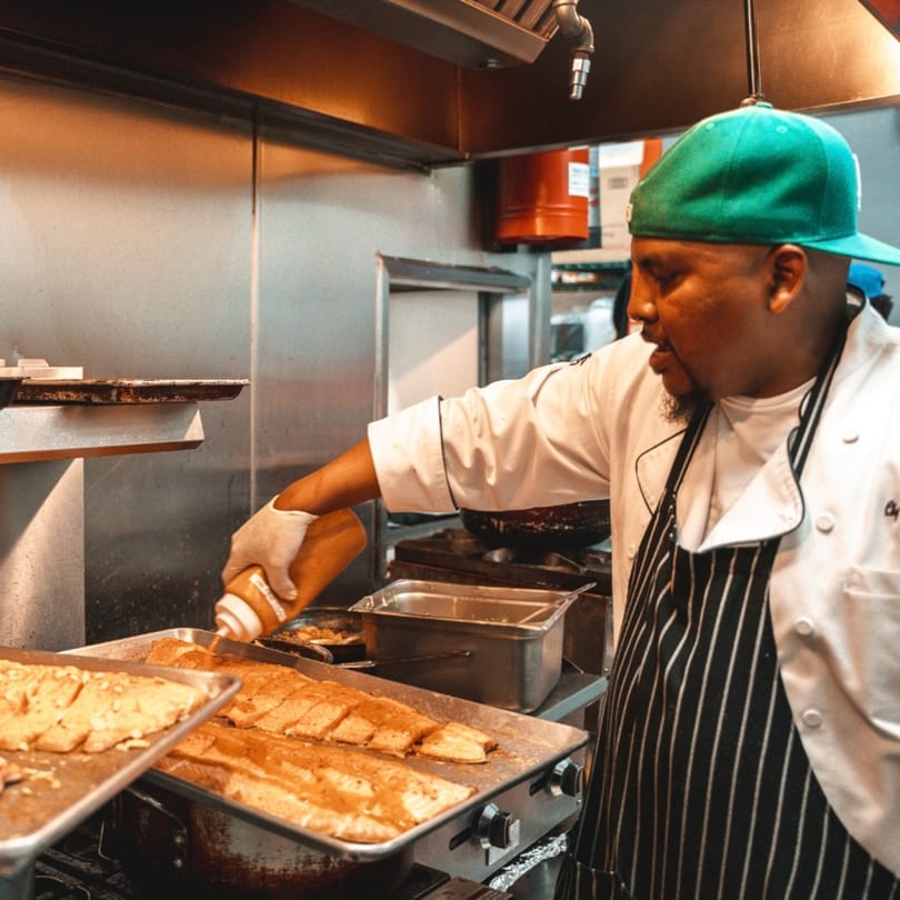A chef in a green hat and striped apron drizzles sauce over Caribbean fish fillets on a tray.