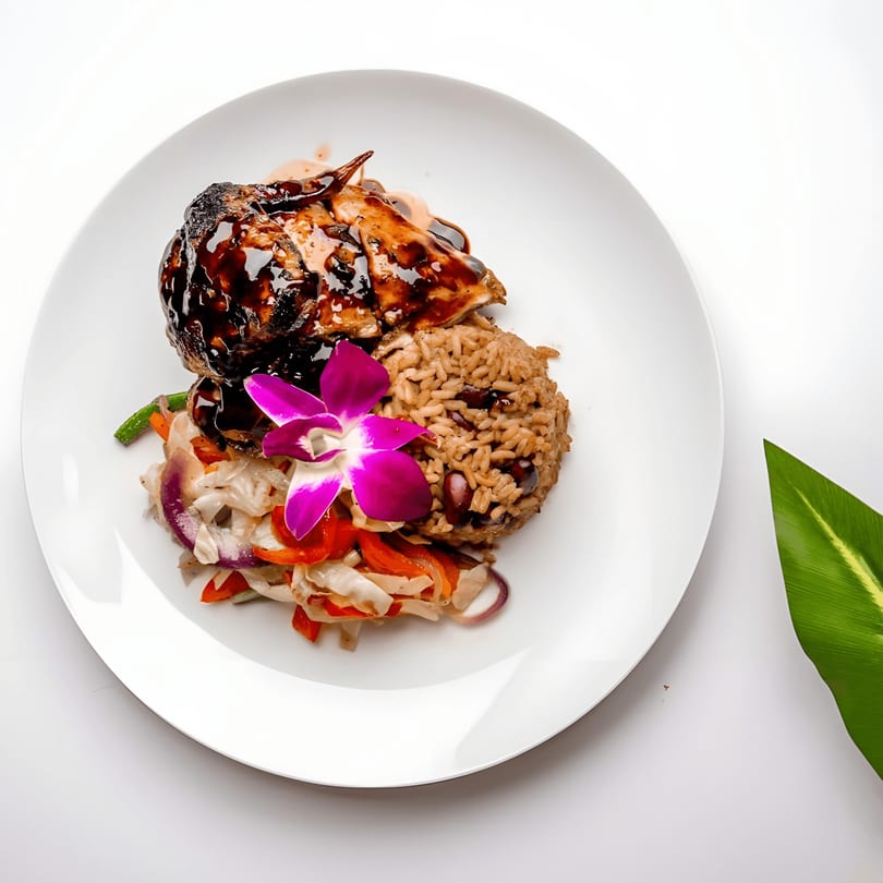 Caribbean jerk chicken with rice and peas, sautéed vegetables, and an orchid garnish on a white plate.